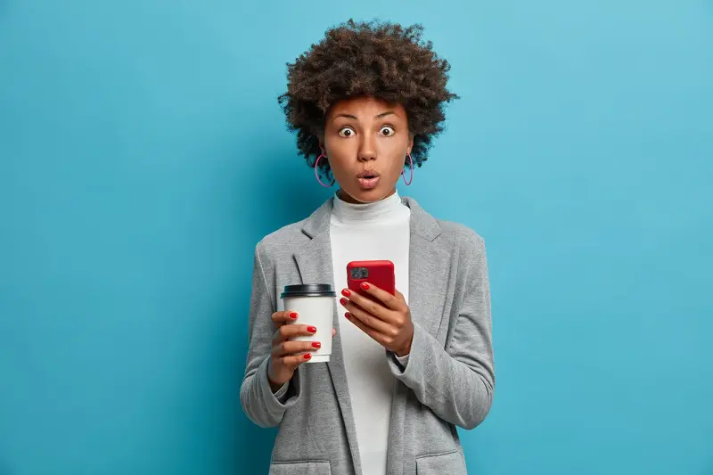 A surprised woman holds a phone and a coffee cup against a blue wall.