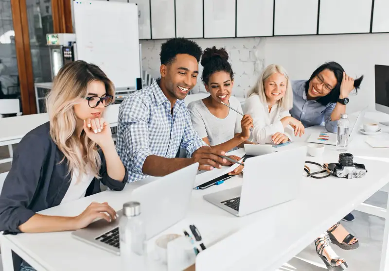 A diverse team laughs while working on laptops around a white table.