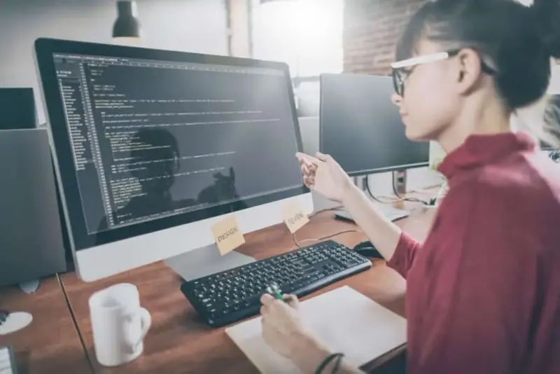 A woman in glasses writes code on a desktop computer at a wooden desk.